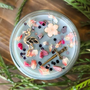 May include: A blue candle with a flamingo, a sword, and pink flower charms. The candle is in a glass jar and has a blue wax base with pink and black glitter.