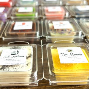 May include: A close-up of two wax melts in clear plastic containers. The wax melt on the left is white with black specks and says "Dark Academia" with the scent "Marigold & Leather". The wax melt on the right is yellow and says "Bee Happy" with the scent "Honey & Rose".