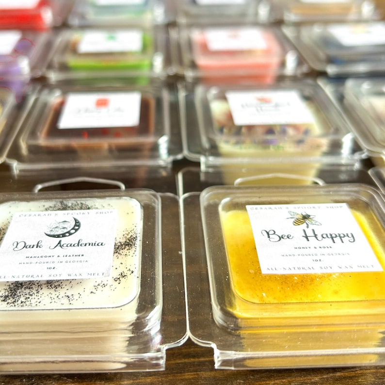 May include: A close-up of two wax melts in clear plastic containers. The wax melt on the left is white with black specks and says "Dark Academia" with the scent "Marigold & Leather". The wax melt on the right is yellow and says "Bee Happy" with the scent "Honey & Rose".