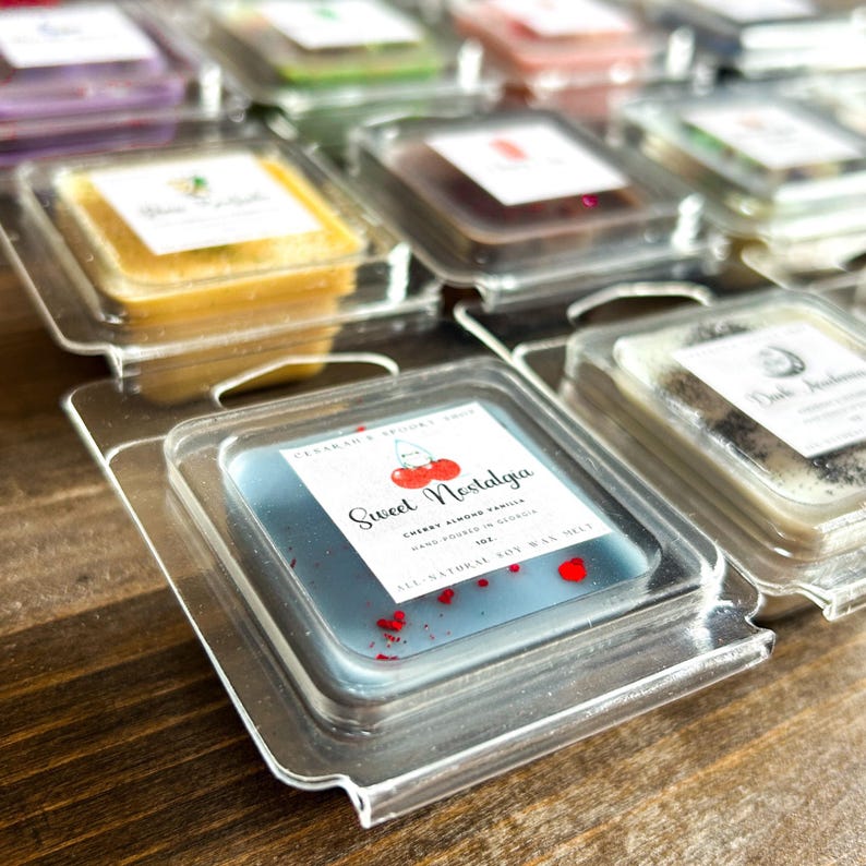 May include: A close-up of a blue soy wax melt with red flecks. The wax melt is in a clear plastic container and has a label that reads "Sweet Nostalgia Cherry Almond Vanilla Hand Poured in Georgia 1 oz All Natural Soy Wax Melt".