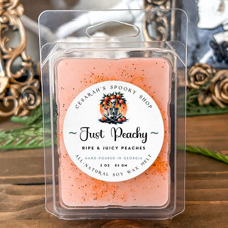 May include: A peach-colored soy wax melt in a clear plastic container. The label reads "Just Peachy" with "Ripe & Juicy Peaches" below. The label also includes a skeleton graphic and states "Hand-poured in Georgia" and "3 oz 85 gm".