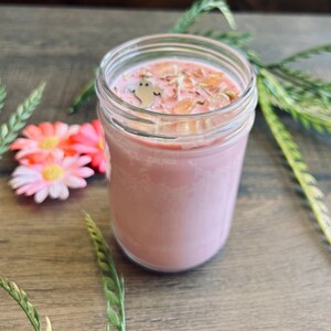 May include: A pink candle in a clear glass jar with a silver ghost-shaped charm and small orange crystal-like pieces embedded in the wax surface. The candle is filled with pink wax and features a smooth, cylindrical glass container design.