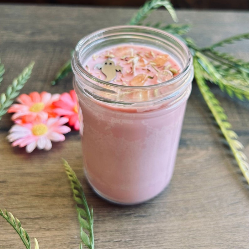 May include: A pink candle in a clear glass jar with a silver ghost-shaped charm and small orange crystal-like pieces embedded in the wax surface. The candle is filled with pink wax and features a smooth, cylindrical glass container design.