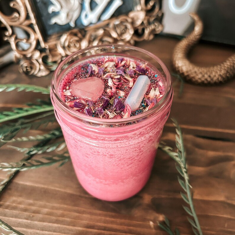 May include: A pink candle in a glass jar topped with dried flower petals, a heart-shaped pink stone, and a small white cylindrical stone. The wax surface is covered in fine glitter, and the jar has a textured, frosted appearance on the exterior.