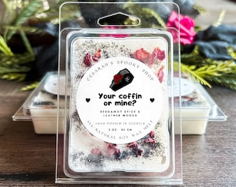 May include: A white soy wax melt in a clear plastic container with a black and white label that says "CESARAH'S SPOOKY SHOP Your coffin or mine? BERGAMOT SPICE & LEATHER WOODS HAND-POURED IN GEORGIA ALL-NATURAL SOY WAX MELT 3 OZ 85 GM". The wax melt is decorated with dried rose petals and black glitter.