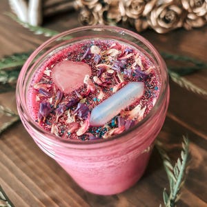 May include: A pink candle in a glass jar topped with a heart-shaped pink stone, a white crystal point, scattered dried purple and beige flower petals, and fine glitter. The wax surface is vibrant pink, and the jar has a clear glass rim.