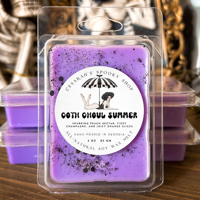 May include: A purple soy wax melt in a clear plastic clamshell. The label reads "Goth Ghoul Summer" with a graphic of a woman under an umbrella. The scent is described as sparkling peach nectar, fizzy champagne, and juicy orange slices. It weighs 3 oz (85 g).
