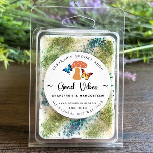 May include: A rectangular wax melt in a clear plastic clamshell. The wax is off-white with green and blue glitter. A circular label reads "Good Vibes" and "Grapefruit & Mangosteen". The label also says "Hand-poured in Georgia" and "3 oz 85 gm".