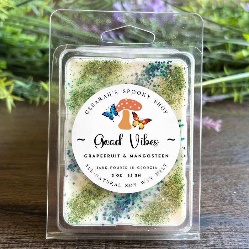 May include: A rectangular wax melt in a clear plastic clamshell. The wax is off-white with green and blue glitter. A circular label reads "Good Vibes" and "Grapefruit & Mangosteen". The label also says "Hand-poured in Georgia" and "3 oz 85 gm".