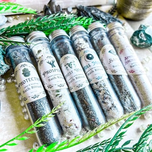 May include: Six small glass tubes filled with white and green bath salts. Each tube has a label with a different name and description, such as "Protection", "Prosper", "Forest Witch", "Full Moon", "Salt", and "Aphrodite Enchantress".