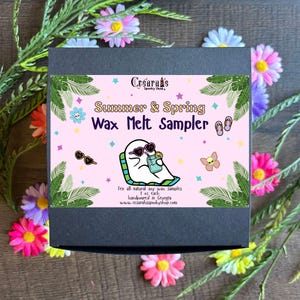 May include: A black box with a pink label that says "Cesaran's Spooky Shop Summer & Spring Wax Melt Sampler". The label features a cartoon ghost wearing sunglasses and holding a glass of lemonade. The text on the label says "Ten all-natural soy wax samples 1 oz each handpoured in Georgia www.cesarahsspookyshop.com".