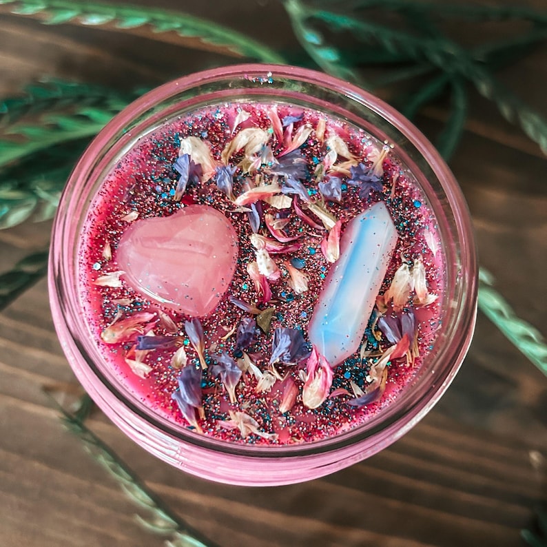 May include: A round candle in a glass jar with pink wax topped with blue and pink glitter, dried flower petals, a smooth pink heart-shaped stone, and a translucent white crystal point. The surface is decorated with scattered botanical elements.