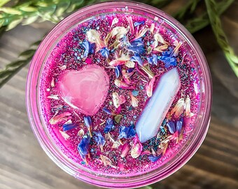 May include: A pink candle in a clear glass jar, decorated with a pink heart-shaped stone, a clear crystal, blue flower petals, and glitter. The candle is surrounded by green foliage.