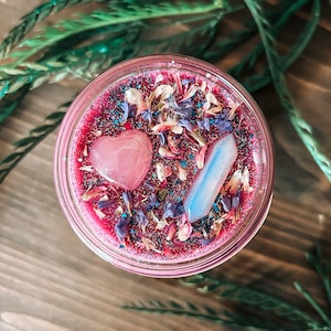 May include: A round candle in a glass jar with deep pink wax topped with blue and white dried flower petals, iridescent glitter, a smooth pink heart-shaped stone, and a translucent, pale blue, pointed crystal pillar embedded in the surface.