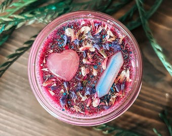 May include: A round candle in a glass jar with deep pink wax topped with blue and white dried flower petals, iridescent glitter, a smooth pink heart-shaped stone, and a translucent, pale blue, pointed crystal pillar embedded in the surface.