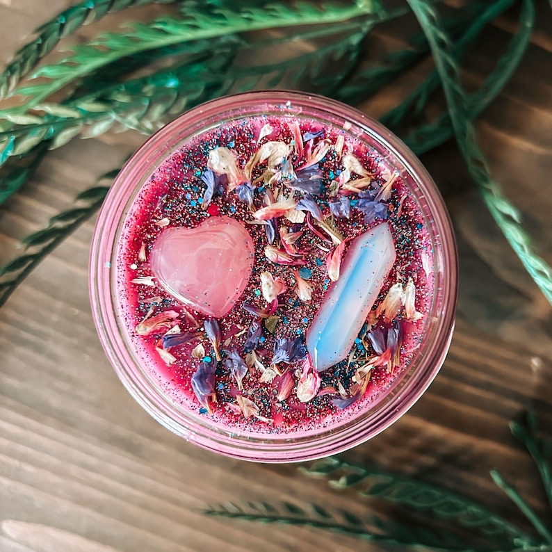 May include: A round candle in a glass jar with deep pink wax topped with blue and white dried flower petals, iridescent glitter, a smooth pink heart-shaped stone, and a translucent, pale blue, pointed crystal pillar embedded in the surface.