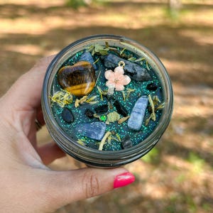 May include: A glass jar candle filled with dark green wax, topped with green glitter, dried herbs, a heart-shaped tiger's eye stone, several blue and black crystals, and a small, light pink flower-shaped charm attached to the candle wick.