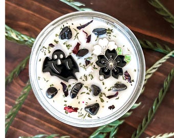 May include: A round white candle in a glass jar topped with scattered dark stones, dried herbs, and two metal charms. One charm is a black ghost figure, and the other is a black and white flower blossom with a small green leaf accent.