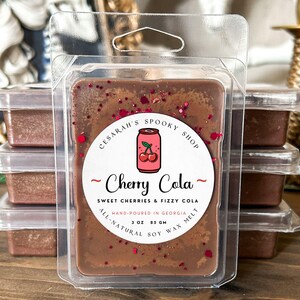 May include: A brown wax melt with red glitter, packaged in a clear plastic container. The label reads "Cherry Cola" with a cherry soda can illustration. It is hand-poured in Georgia and contains 3 oz (85 g) of all-natural soy wax.