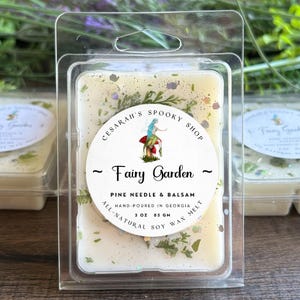 May include: A clear plastic clamshell package containing a soy wax melt. The wax melt is off-white with green and glittery accents. The label reads "Fairy Garden" with a fairy illustration and "Pine Needle & Balsam". It is hand-poured in Georgia, 3 oz (85 g).
