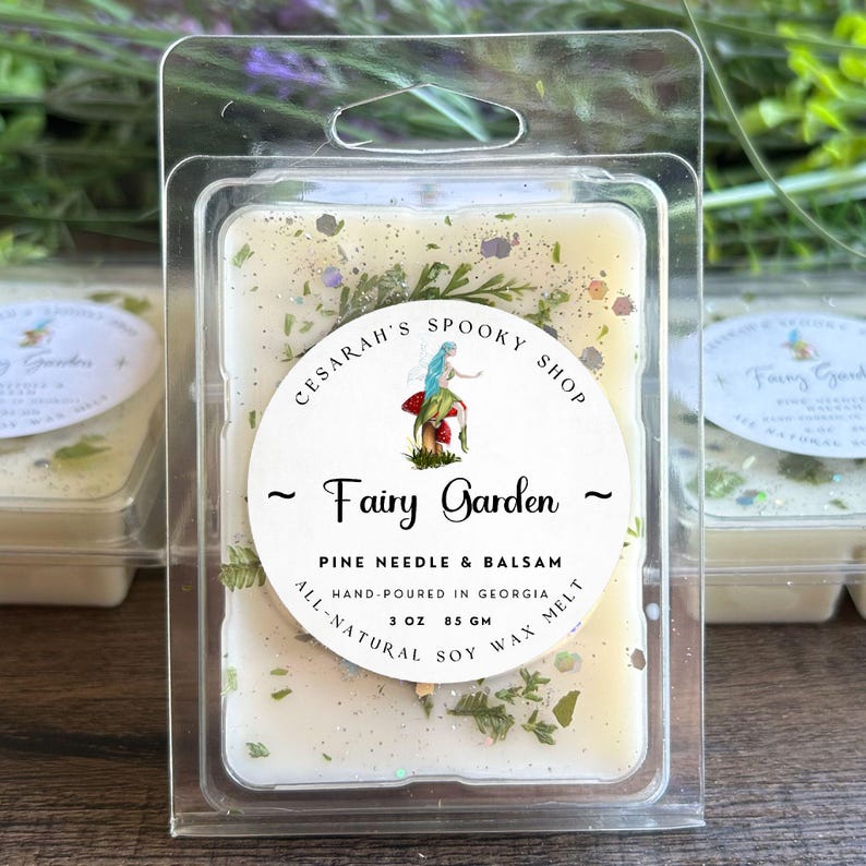 May include: A clear plastic clamshell package containing a soy wax melt. The wax melt is off-white with green and glittery accents. The label reads "Fairy Garden" with a fairy illustration and "Pine Needle & Balsam". It is hand-poured in Georgia, 3 oz (85 g).