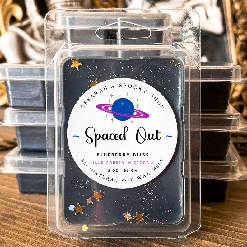 May include: A dark blue wax melt in a clear plastic clamshell. The label reads "Spaced Out - Blueberry Bliss" with a planet graphic. It is hand-poured in Georgia and contains 3 oz (85 gm) of all-natural soy wax melt. Gold star-shaped glitter is sprinkled throughout.