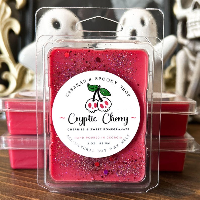 May include: A red wax melt in a clear plastic container. The label reads "Cryptic Cherry" with a cherry skull graphic. The wax melt is 3 oz (85 g) and made with all-natural soy wax. The text "Cesarah's Spooky Shop" is also visible.