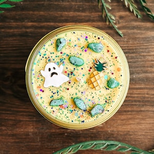 May include: A round yellow candle in a gold metal tin, topped with multicoloured glitter, several small green stone-like pieces, a white ghost-shaped metal charm with a sad face, and a yellow and green pineapple-shaped metal charm.