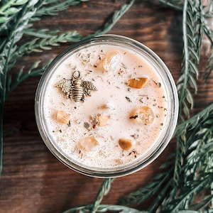 May include: A round glass candle container filled with light beige wax, topped with a gold-toned metal bee charm, several small translucent yellow crystals, and scattered dried botanical herbs across the surface of the candle wax.