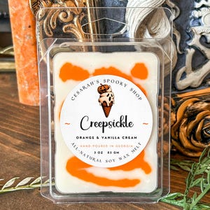 May include: A white soy wax melt with orange swirls, packaged in a clear plastic container. The label reads "Creepsickle" with an illustration of an ice cream cone. The text also says "Orange & Vanilla Cream" and "Hand-poured in Georgia, 3 oz".