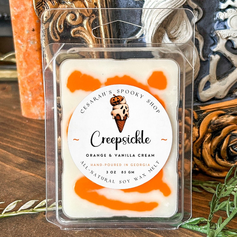 May include: A white soy wax melt with orange swirls, packaged in a clear plastic container. The label reads "Creepsickle" with an illustration of an ice cream cone. The text also says "Orange & Vanilla Cream" and "Hand-poured in Georgia, 3 oz".
