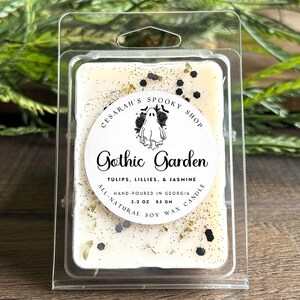 May include: A soy wax candle melt in a clear plastic container. The label reads "Gothic Garden" with a ghost graphic. The candle is off-white with decorative specks and the text "Tulips, Lillies, & Jasmine". It is hand-poured in Georgia and weighs 3.2 oz.