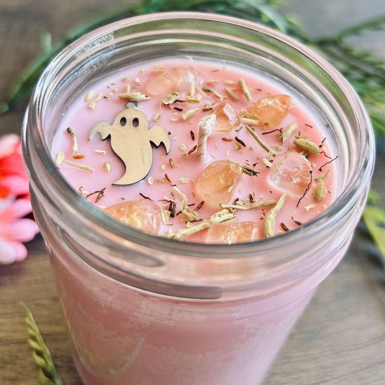 May include: A pink candle in a clear glass jar featuring a silver ghost-shaped charm, several translucent orange crystal stones, and scattered dried herbs on the wax surface. A single cotton wick is centered among the decorative elements.