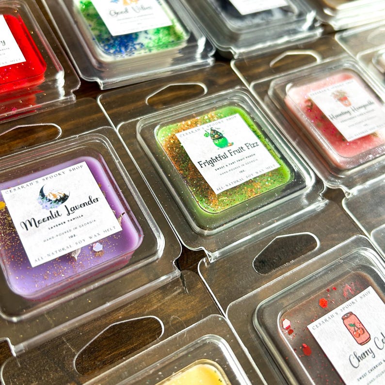 May include: Assortment of small square wax melts in clear plastic containers. The wax melts are various colors and have glitter and text on top. The text includes "Moonlit Lavender", "Frightful Fruit Fizz", and "Cherry Cola".