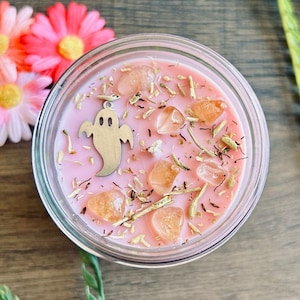 May include: A pink wax candle in a glass jar topped with scattered dried herbs, several translucent orange tumbled stones, and a silver-toned metal ghost charm. The candle surface is smooth and light pink, with a single visible wick center.