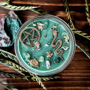May include: A round glass candle container filled with sage green wax, topped with a bronze-colored metal pentagram charm, a bronze-colored metal snake charm, several small light green tumbled stones, and scattered dried botanical fragments.