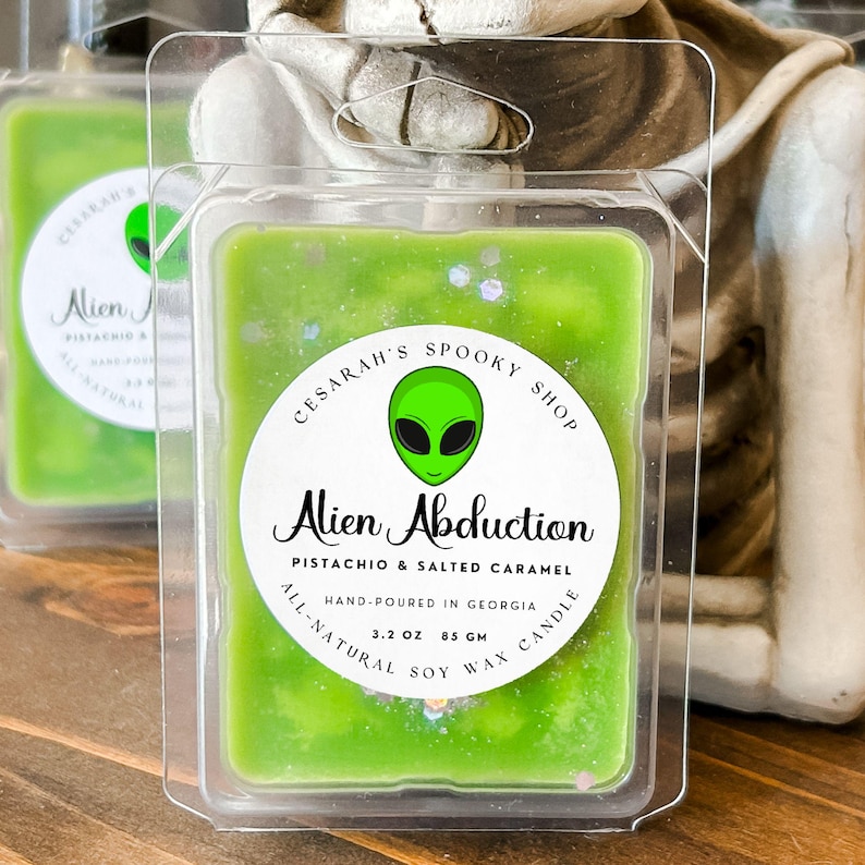 May include: A green soy wax candle in a plastic container. The label reads "Alien Abduction" with the scent "Pistachio & Salted Caramel." The label also includes the text "Hand-poured in Georgia" and "3.2 OZ 85 GM."