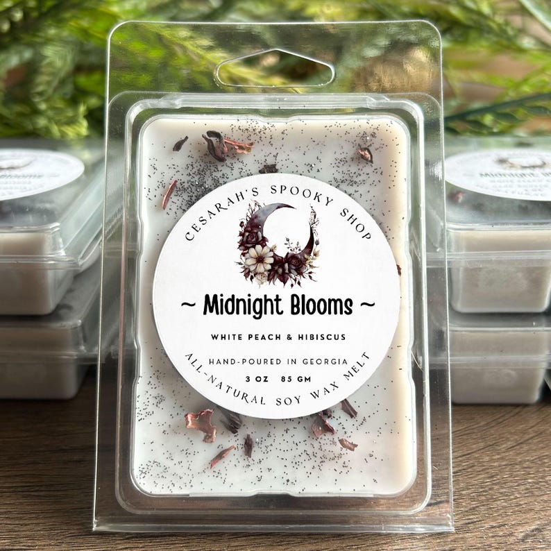 May include: A white wax melt with the text "Midnight Blooms" and "White Peach & Hibiscus." The label also reads "Hand-poured in Georgia" and "3 oz 85 gm." The wax melt is in a clear plastic container.