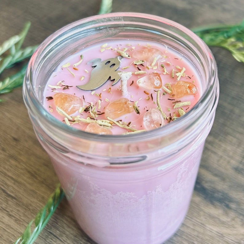 May include: A pink candle in a clear glass jar, topped with a silver ghost-shaped charm, several small orange-tinted crystals, and dried herbs surrounding a central white wick. The wax is smooth and light pink, filling the glass container.