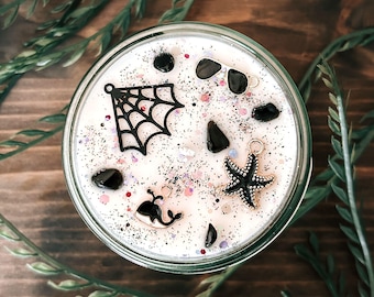 May include: A white candle in a glass jar topped with black glitter and various charms including a black spiderweb, a black starfish, a small whale, and black stones. The surface is decorated with scattered black and pink glitter particles.