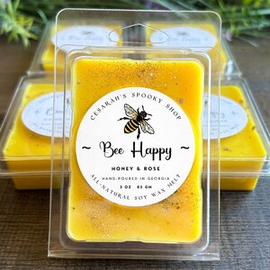 May include: Yellow soy wax melts in clear plastic packaging. The label reads "Bee Happy" with a bee illustration, and "Honey & Rose." Each melt is 3 oz (85 g). The text also says "Hand-poured in Georgia."