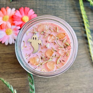 May include: A round glass candle container filled with pink wax, topped with scattered dried herbs, several smooth orange-toned crystals, and a silver-colored metal ghost charm. The candle is viewed from directly above.