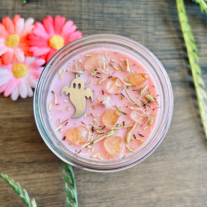 May include: A round glass candle container filled with pink wax, topped with scattered dried herbs, several smooth orange-toned crystals, and a silver-colored metal ghost charm. The candle is viewed from directly above.