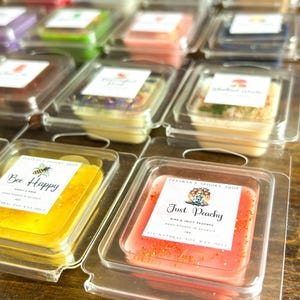 May include: Close-up of two small, square wax melts in clear plastic containers. The wax melt on the left is yellow and says "Bee Happy" with a bee illustration. The wax melt on the right is pink and says "Just Peachy" with a peach illustration.