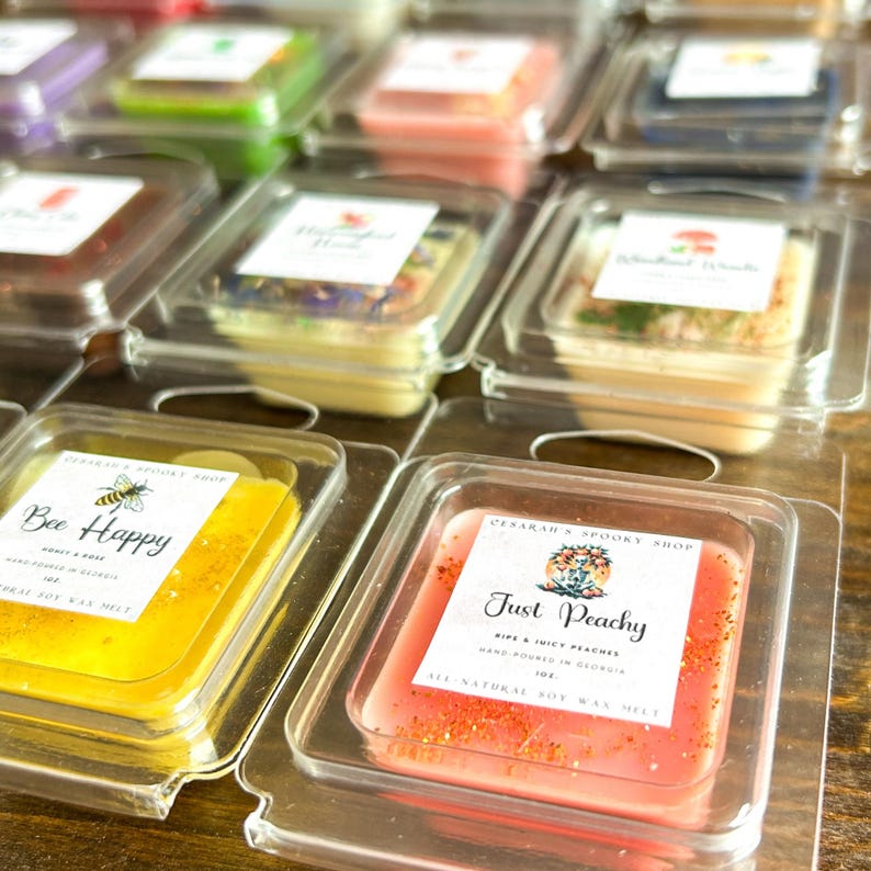 May include: Close-up of two small, square wax melts in clear plastic containers. The wax melt on the left is yellow and says "Bee Happy" with a bee illustration. The wax melt on the right is pink and says "Just Peachy" with a peach illustration.