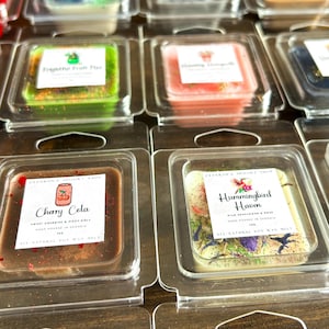 May include: A close-up of ten small square wax melts in clear plastic containers. The wax melts are scented and have different designs and colors. The scents are "Cherry Cela", "Hummingbird Haven", "Grapefruit Fruit Punch", and "Honeycomb".