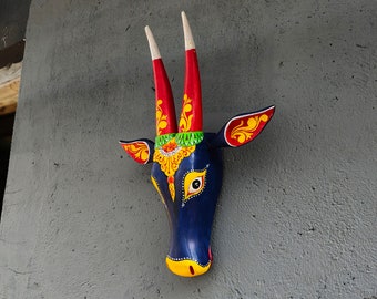 Pode incluir: Uma máscara de animal de madeira azul com detalhes pintados em vermelho, amarelo e verde. A máscara tem dois chifres longos e um rosto estilizado com um único olho.