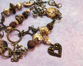 May include: A brown and gold necklace with a heart pendant. The necklace is made of metal, beads, and buttons. The beads are brown, gold, and white. The buttons are brown and white.