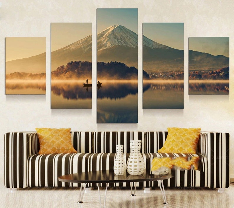 Shows Mount Fuji.  Five-piece canvas art print showing a tranquil mountain Fuji  scene with a snow-covered summit reflected in still water. A boat with people is on the lake. The artwork is above a striped sofa with yellow cushions.