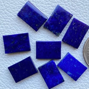 Puede incluir: Siete piedras preciosas rectangulares de lapislázuli, cada una con un color azul intenso y motas de pirita dorada. Las piedras están dispuestas sobre una superficie blanca.